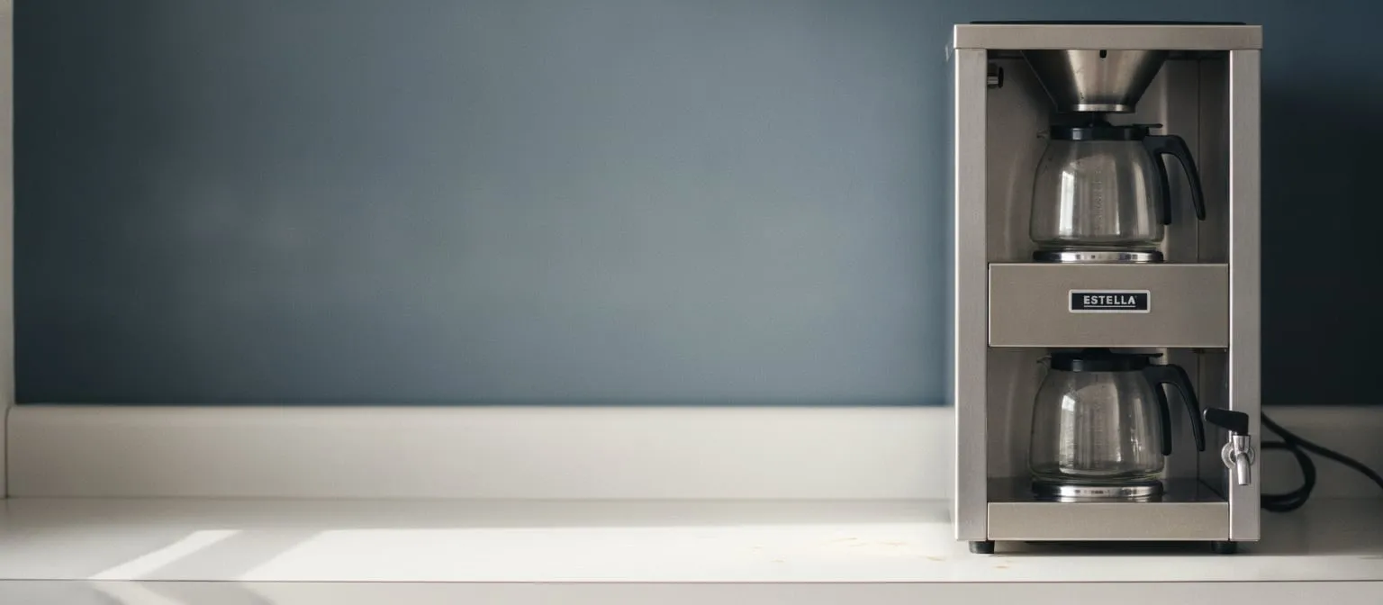 Stainless steel commercial batch coffee brewer with two glass decanters on warming plates, standing on a break-room counter